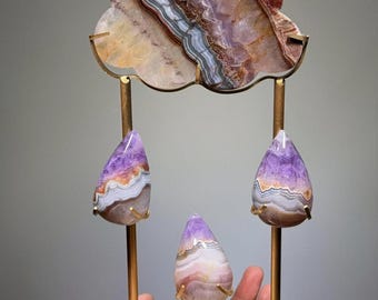 May include: A decorative display stand featuring a cloud-shaped slice of banded agate in shades of purple, brown, and white, with three matching teardrop-shaped amethyst and agate stones suspended on gold-toned metal rods and a square base.