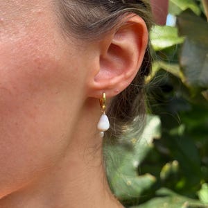 Puede incluir: Un pendiente de aro dorado con un colgante blanco en forma de lágrima y una pequeña cuenta blanca. El pendiente se lleva en la oreja, con un fondo verde borroso.