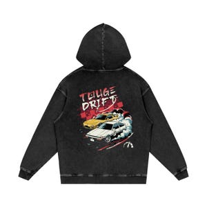 Op de afbeelding: Zwarte hoodie met een afbeelding van twee driftende auto's en de woorden "TOUGE DRIFT" in rood en wit. De auto's zijn geel en wit, met rookeffecten. De hoodie heeft een vintage wassing.