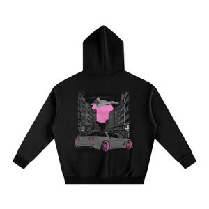 Op de afbeelding: Zwarte hoodie met een grafische print op de achterkant. Het ontwerp omvat een roze shirt, een straalvliegtuig en een grijze sportwagen met roze wielen, tegen een stadsgezicht.
