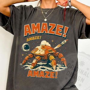 May include: A charcoal gray short-sleeve t-shirt featuring a graphic of an orange robotic creature on a moon surface. The text AMAZE! appears in orange above and below the robot. A small rocket and astronaut helmet are also visible in the design.
