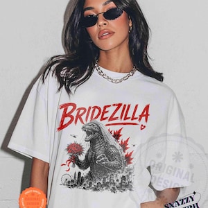 May include: White t-shirt with a graphic of Godzilla holding a bouquet of flowers, with the word "Bridezilla" in red. The design includes a cityscape and maple leaves. The shirt has a round neck and short sleeves.