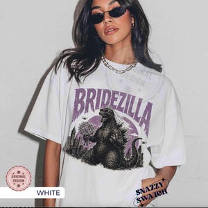 May include: White t-shirt with a purple graphic that says "Bridezilla" above an illustration of Godzilla holding a bouquet. The design includes a cityscape and a sunset. The shirt has short sleeves.
