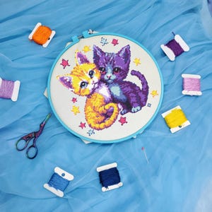 May include: A cross-stitch embroidery hoop featuring two adorable kittens, one orange and one purple, surrounded by colorful thread spools, scissors, and a pin. The kittens are hugging each other, with star accents.