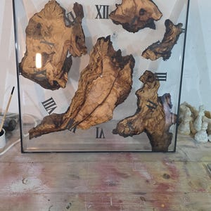 May include: Square wall clock with a clear resin face, featuring natural wood pieces and black Roman numerals. The clock's wooden elements have a rich, brown colour, and the frame is black.
