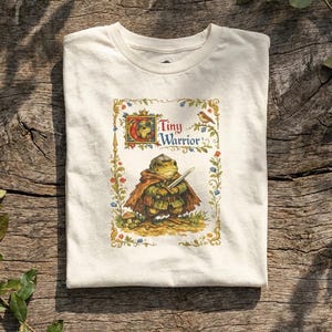 May include: Off-white t-shirt featuring a whimsical illustration of a frog warrior with a sword, wearing a cape. The design includes the text "Tiny Warrior" in a decorative frame with floral and bird accents. The shirt is folded on a wooden surface.