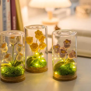 May include: Three glass cylinders with crochet flowers and fairy lights. The flowers are yellow, white, and lavender. The cylinders sit on cork bases and contain green moss. The lights add a warm glow.
