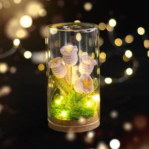 May include: A glass terrarium with a cork base, filled with crocheted lavender tulips and green moss. Tiny fairy lights illuminate the scene, creating a warm glow. The background is dark with bokeh lights.