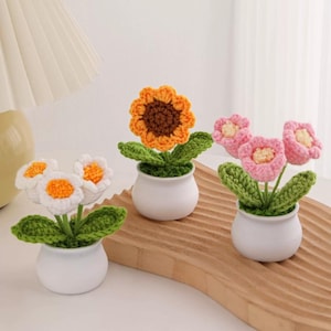 May include: Three crochet flower arrangements in white ceramic pots. One features a sunflower with orange and brown petals, the others have white and orange, and pink and cream flowers. Green leaves and stems complete the handmade look.
