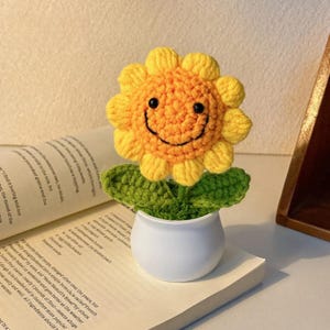 以下が含まれることがあります： 笑顔の顔、黄色の花びら、緑の葉を持つかぎ針編みのヒマワリが、小さな白い鉢に入っています。手作りの花は本の上に置かれ、陽気な装飾アクセントになっています。