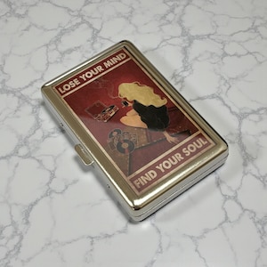 May include: A silver metal cigarette case with a red and brown graphic. The graphic features a woman with blonde hair, a record player, and the text "LOSE YOUR MIND FIND YOUR SOUL".