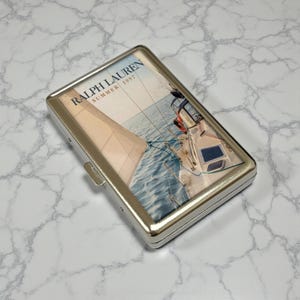 May include: A silver metal cigarette case with a nautical image. The image features a sailboat on the water with the text "RALPH LAUREN SUMMER 1997". The case has a rectangular shape and a clasp closure.