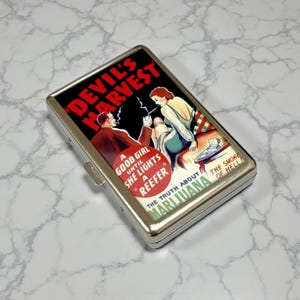 Puede incluir: Un estuche de cigarrillos de metal plateado con un diseño de cartel de película vintage. El cartel presenta el texto "DEVIL'S HARVEST" en rojo y las palabras "A GOOD GIRL UNTIL SHE LIGHTS A REEFER". El estuche también tiene las palabras "MARIJUANA" y "THE SMOKE OF HELL".