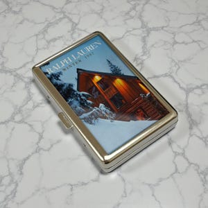 Könnte beinhalten: Eine silberne Zigarettenetui aus Metall mit rechteckigem Design. Die Hülle zeigt eine Winterszene mit einer Hütte und dem Text "RALPH LAUREN WINTER 1997" auf der Vorderseite. Die Hülle wird mit einem Metallverschluss geschlossen.