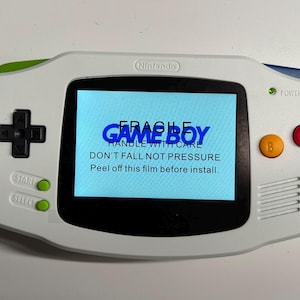 May include: A white handheld video game console with a black directional pad, a yellow B button, and a red A button. The screen displays the text: FRAGILE GAME BOY HANDLE WITH CARE DON'T FALL NOT PRESSURE Peel off this film before install.