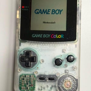 以下が含まれることがあります： GAME BOYおよびNintendoの文字が表示された画面を持つ、透明なハンドヘルド型ビデオゲーム機。黒い十字キー、AボタンとBボタン、SelectボタンとStartボタン、そして内部の回路基板が見えるデザインとなっています。