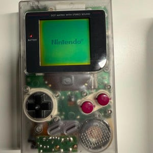 May include: A handheld video game console with a transparent plastic shell revealing internal green circuitry. It features a black screen bezel with the text DOT MATRIX WITH STEREO SOUND and Nintendo, a black directional pad, two magenta buttons, and a speaker.