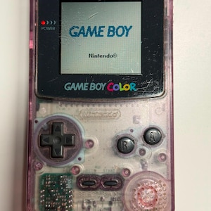 Peut inclure: Une console de jeu vidéo portable violette translucide avec un écran affichant le texte GAME BOY et Nintendo. L'appareil dispose d'une croix directionnelle, des boutons A et B, des boutons Select et Start, et un haut-parleur visible.