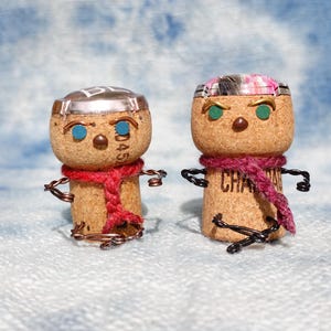 以下が含まれることがあります： ワイヤー製の腕と脚、赤またはピンクのスカーフをつけた、2つの気まぐれなコルクのフィギュア。片方は銀色のホイルハットと青い目を、もう片方は模様入りの帽子と緑の目をしています。コルクには「CHAMPAGNE」の文字と「045」の数字が飾られています。