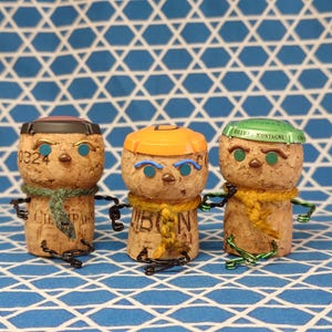 以下が含まれることがあります： ボトルキャップの帽子、針金の手足、スカーフをつけた、気まぐれなコルクのフィギュア3体。コルクには青い目のペイントが施されています。コルクの1つには「0324」の数字がプリントされています。青と白の模様の背景に立っています。