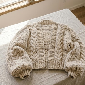 Könnte beinhalten: Beige Strickjacke mit Zopfmuster. Der offene Cardigan hat lange, voluminöse Ärmel und gerippte Bündchen und Saum. Das grob gestrickte Kleidungsstück wird auf einer neutralen Oberfläche präsentiert. Das Design umfasst ein Mittelteil und Zopfmusterdetails.