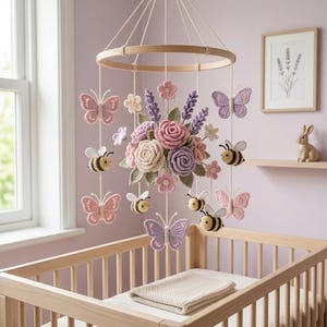 Op de afbeelding: Handgemaakte baby mobile met gehaakte vlinders, bijen en bloemen in pastelroze, paarse en crèmekleuren. De mobile hangt aan een houten ring boven een houten wieg.