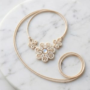 May include: A cream-colored macrame necklace featuring a large flower pendant with a central, round, iridescent stone. The necklace has a delicate, woven design and a long, adjustable cord.