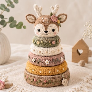 Peut inclure: Un jouet à empiler en crochet avec une tête de cerf surmontée de bois et d'une fleur. Le jouet se compose de cinq anneaux circulaires dans des tons marron, jaune moutarde, rose poudré, crème et vert sauge, chacun orné de petits motifs floraux brodés.