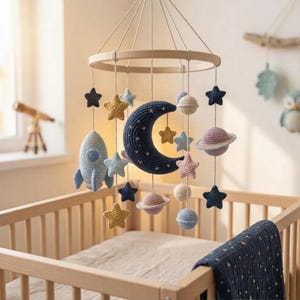 Könnte beinhalten: Handgefertigtes Baby-Mobile mit einer blauen Rakete, einem Mond, Sternen und Planeten. Die Dekoration für das Kinderzimmer hängt an einem Holzring über einem Kinderbett. Das Mobile ist aus weichem, gehäkeltem Garn in Blau-, Gelb- und Rosatönen gefertigt.