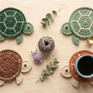 May include: Crocheted turtle-shaped coasters in shades of green, brown, and beige, with a small purple turtle figurine and a ball of yarn. One coaster holds a blue mug with coffee. A gold spoon rests on one coaster.