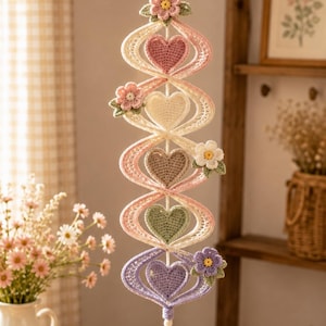 May include: A vertical crochet wall hanging featuring a spiraling structure with five stuffed hearts in shades of pink, cream, beige, sage green, and lavender. The piece is decorated with small crocheted flowers and finished with a white tassel.