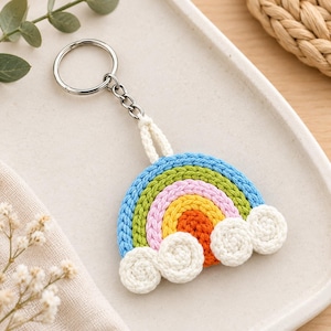 May include: A rainbow keychain crafted from braided cord in vibrant shades of blue, green, pink, yellow, and orange. The arch features three white crocheted clouds at the base and is attached to a silver-toned metal split ring by a short cord loop.