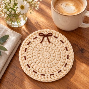 May include: A round, cream-coloured crochet coaster featuring a decorative dark brown stitched border and a small, centred dark brown yarn bow at the top edge. The coaster is crafted from thick yarn with a visible, textured circular stitch pattern.