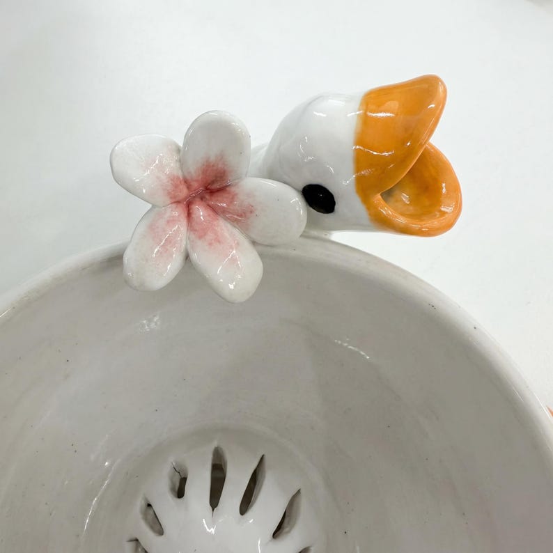May include: A white ceramic bowl featuring a sculpted duck head with an orange beak and a small white flower with a pink centre attached to the rim. The interior of the bowl has a series of vertical slits at the bottom, creating a strainer design.