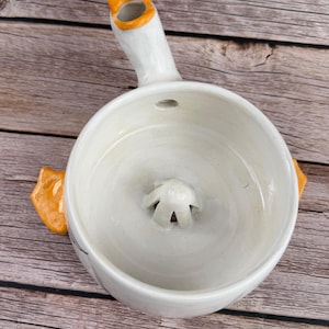 May include: A white ceramic citrus juicer shaped like a duck, featuring a central reamer, an orange beak-shaped spout for pouring, and two orange webbed feet protruding from the sides. The interior bowl is smooth with a glossy white finish.