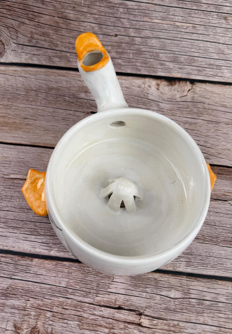 May include: A white ceramic citrus juicer shaped like a duck, featuring a central reamer, an orange beak-shaped spout for pouring, and two orange webbed feet protruding from the sides. The interior bowl is smooth with a glossy white finish.