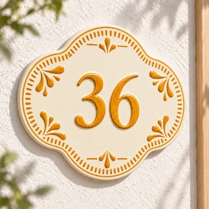 Peut inclure: Une plaque de numéro de maison en céramique crème affichant le nombre 36 dans une police jaune moutarde audacieuse. La plaque présente un bord festonné avec une bordure jaune moutarde et des motifs floraux décoratifs assortis.