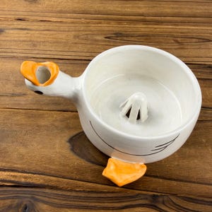 May include: A white ceramic citrus juicer shaped like a duck, featuring an orange beak that serves as a spout, an orange foot at the base, and a central reamer cone inside the bowl. The exterior has simple black line details depicting wings.