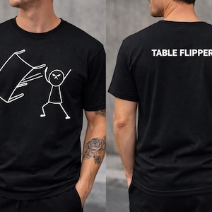 Puede incluir: Camiseta negra con un dibujo blanco de un muñeco de palo volcando una mesa. La parte trasera de la camiseta tiene el texto "TABLE FLIPPER" en blanco. Un diseño divertido para uso diario.