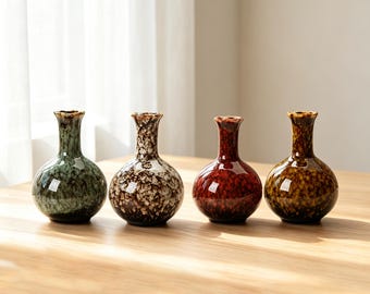 Pode incluir: Quatro pequenos vasos de cerâmica com base arredondada e gargalo estreito, cada um com um acabamento de esmalte brilhante distinto. As cores incluem verde sálvia, um padrão salpicado de branco e castanho, vermelho profundo e âmbar dourado.