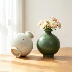 以下が含まれることがあります： 細い首を持つ2つの丸い陶器製の花瓶。1つは光沢のあるオフホワイトで暗い斑点があり、横向きに置かれています。もう1つは無地のマットなフォレストグリーンで、緑の茎を持つ淡いピンクの花束が生けられています。