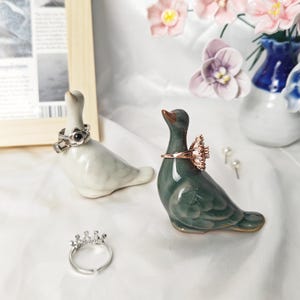May include: Two ceramic bird-shaped ring holders, one light gray and one dark green, each displaying a ring on its neck. A silver crown-shaped ring rests on the surface, and two small stud earrings are positioned near the green bird figure.
