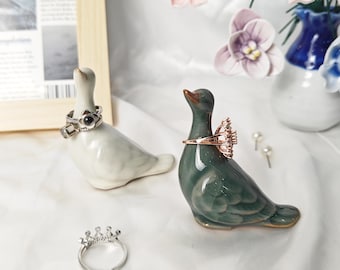 May include: Two ceramic bird-shaped ring holders, one light gray and one dark green, each displaying a ring on its neck. A silver crown-shaped ring rests on the surface, and two small stud earrings are positioned near the green bird figure.
