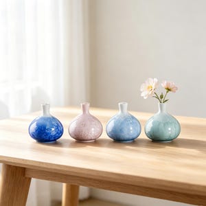 May include: Four small ceramic vases in blue, pink, and green sit on a wooden table. One vase holds delicate pink and white flowers. The vases have a rounded shape with a narrow neck. The table is made of wood.