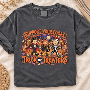 May include: A charcoal gray short-sleeve t-shirt featuring a colorful graphic of three children in Halloween costumes with the text "SUPPORT YOUR LOCAL TRICK OR TREATERS" in orange. The design includes a vampire, a cat, and a skeleton character.