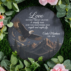 May include: A round black slate memorial plaque featuring a flying eagle, a mountain landscape, and a shooting star. The text reads: Love NEVER TRULY LEAVES it simply rises. NOW YOU SOAR WHERE ONLY angels and eagles fly. Carla Martinez 1950 - 2026.