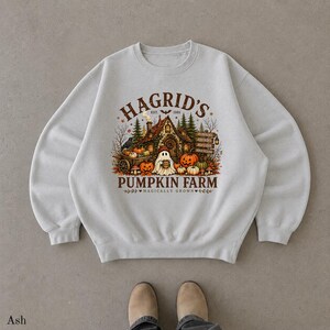 May include: An ash gray crewneck sweatshirt featuring a graphic print of a house with a ghost, pumpkins, and trees. The text reads HAGRID'S PUMPKIN FARM EST 1981 and MAGICALLY GROWN. The design includes a wooden signpost and a small black bird.