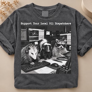 May include: A charcoal grey t-shirt featuring a black and white graphic of an opossum, a skunk, and a raccoon wearing headsets at computer workstations. The text above the graphic reads, Support Your Local 911 Dispatchers, in a white typewriter font.
