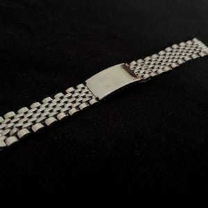 May include: A silver-toned metal watch band featuring a multi-link beads-of-rice style construction. The strap includes a polished rectangular folding clasp mechanism in the center, designed to secure the bracelet around a wrist.