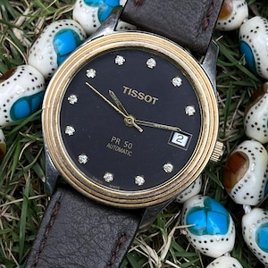 May include: A Tissot PR 50 automatic watch with a black dial, gold-tone bezel, and diamond hour markers. The watch has a brown leather strap and a date window at the 3 o'clock position. The watch is surrounded by white and blue beads.